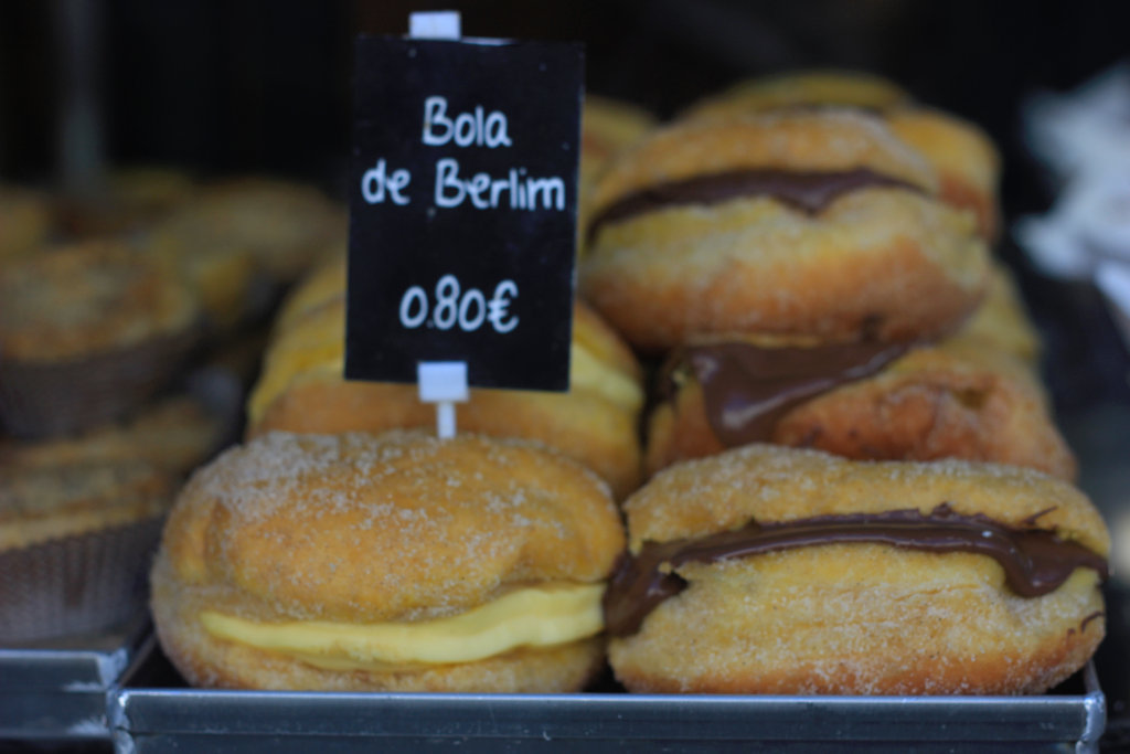 Bolas de berlim num tabuleiro de pastelaria atrás de uma placa manuscrita que diz Bola de Berlim 0,80 euros